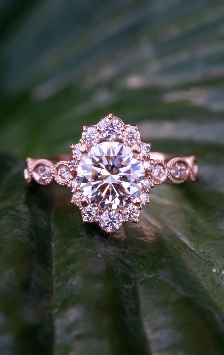 A diamond ring, mounted in rose gold, with a diamond halo and diamonds mounting along the band sits on a large green leaf